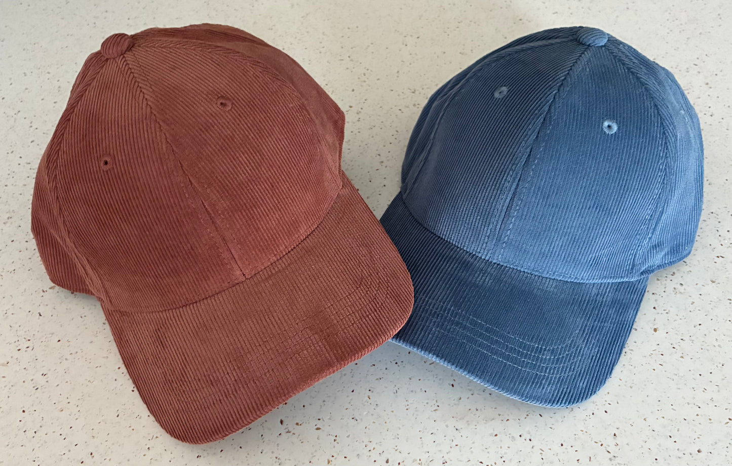 Two organic cotton corduroy caps, side by side against a pale patterned surface. One is Redwood and one Slate Blue.