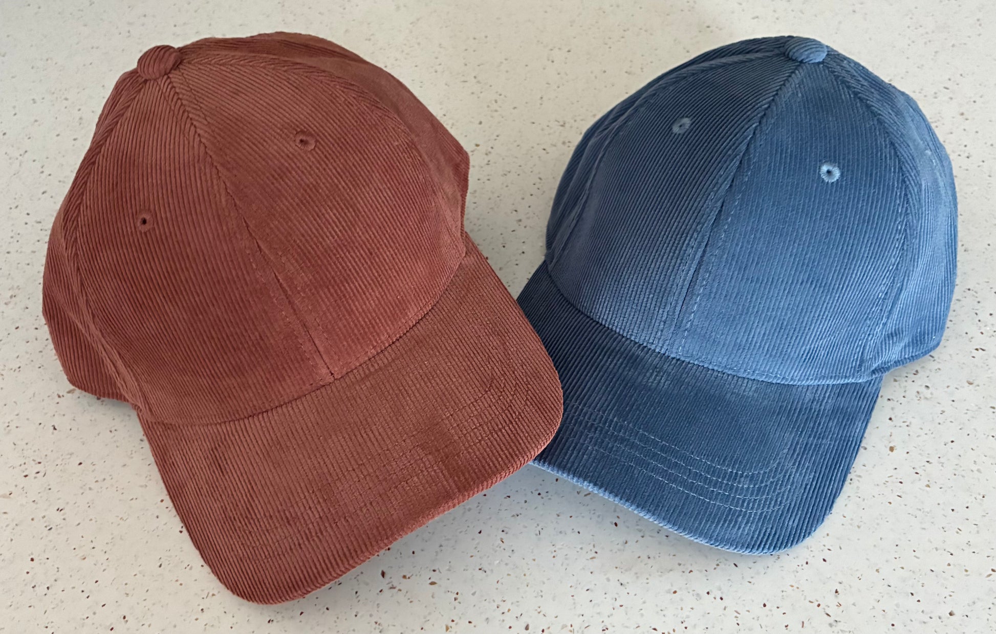 Two organic cotton corduroy caps, side by side against a pale patterned surface. One is Redwood and one Slate Blue.