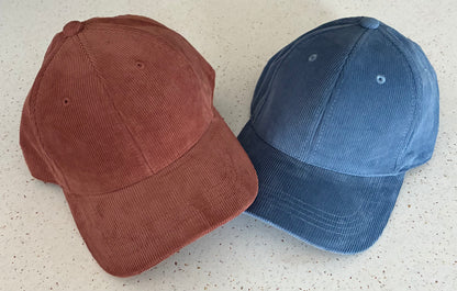 Two organic cotton corduroy caps, side by side against a pale patterned surface. One is Redwood and one Slate Blue.