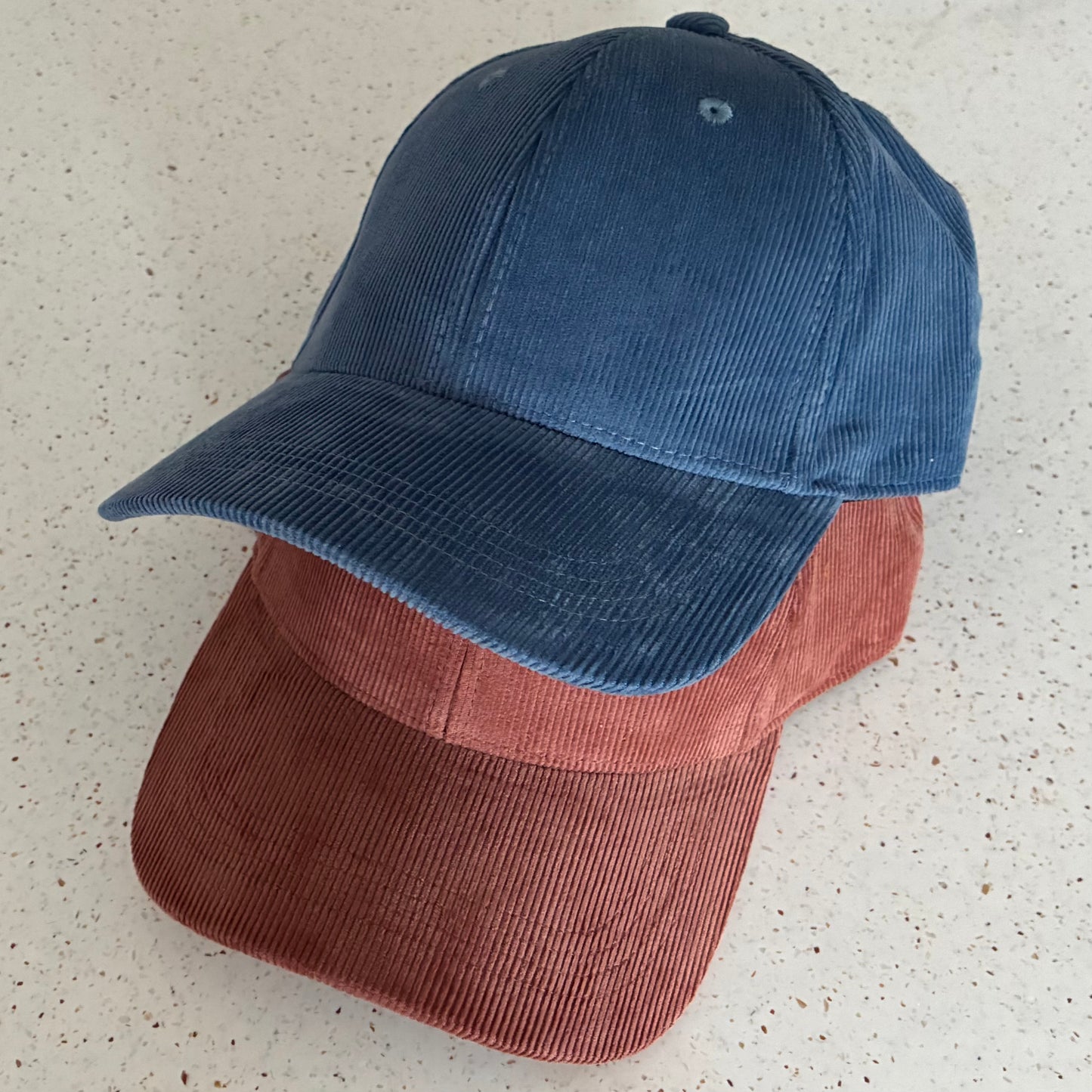 Two corduroy caps, made from 100% organic cotton, stacked on top of each other against a patterned white background. The top cap is a Slate Blue colour, and the bottom one is a rusty colour called Redwood.