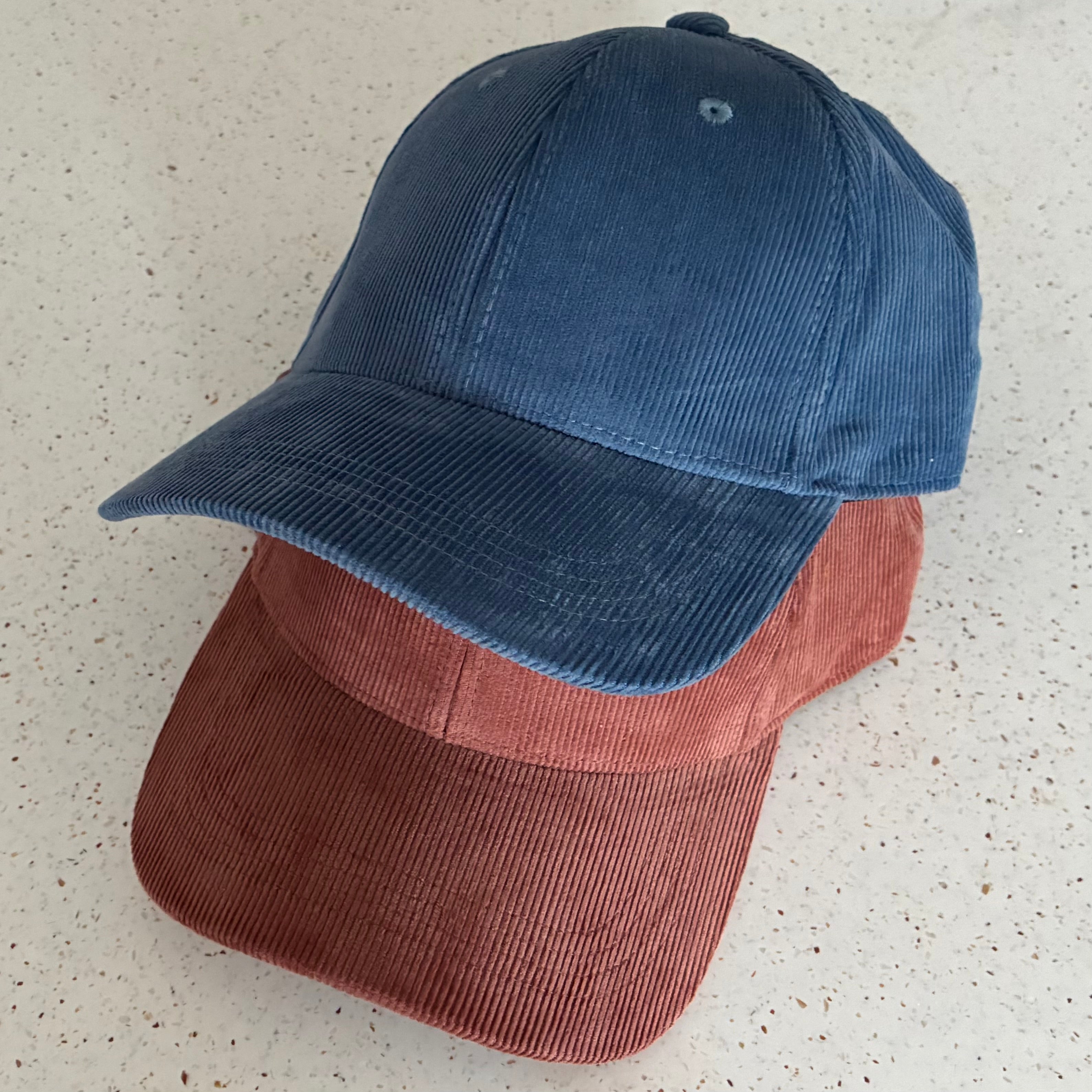 Two corduroy caps, made from 100% organic cotton, stacked on top of each other against a patterned white background. The top cap is a Slate Blue colour, and the bottom one is a rusty colour called Redwood.