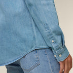 Close-up of the sleeve and cuffs of the light wash blue men's denim shirt also showing the curved back hem against a neutral background