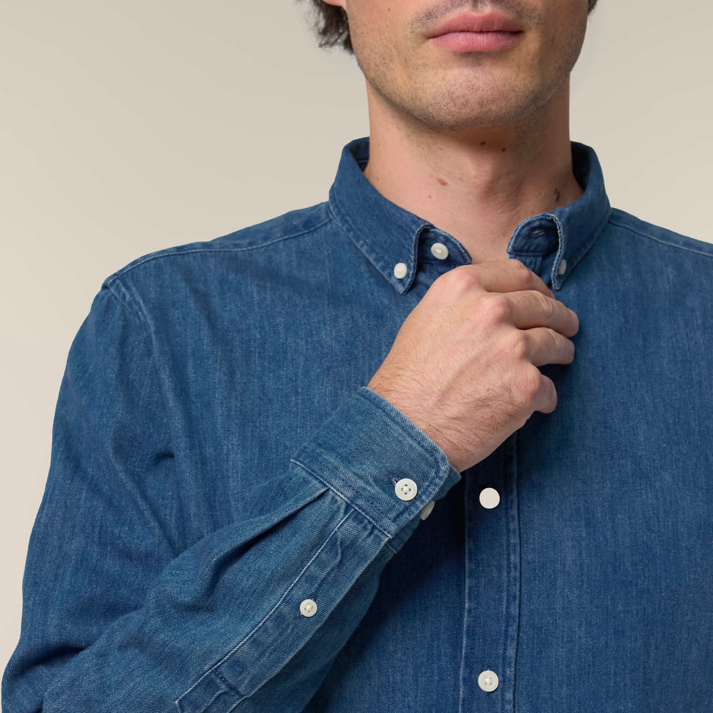Man adjusting the collar of a mid wash blue denim shirt against a neutral background
