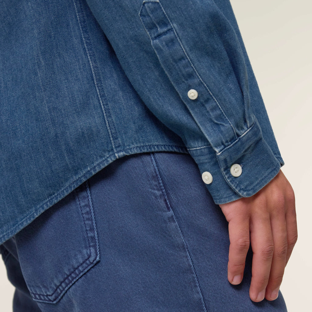 Close-up of the sleeve and cuffs of the mid wash blue men's denim shirt also showing the curved back hem against a neutral background