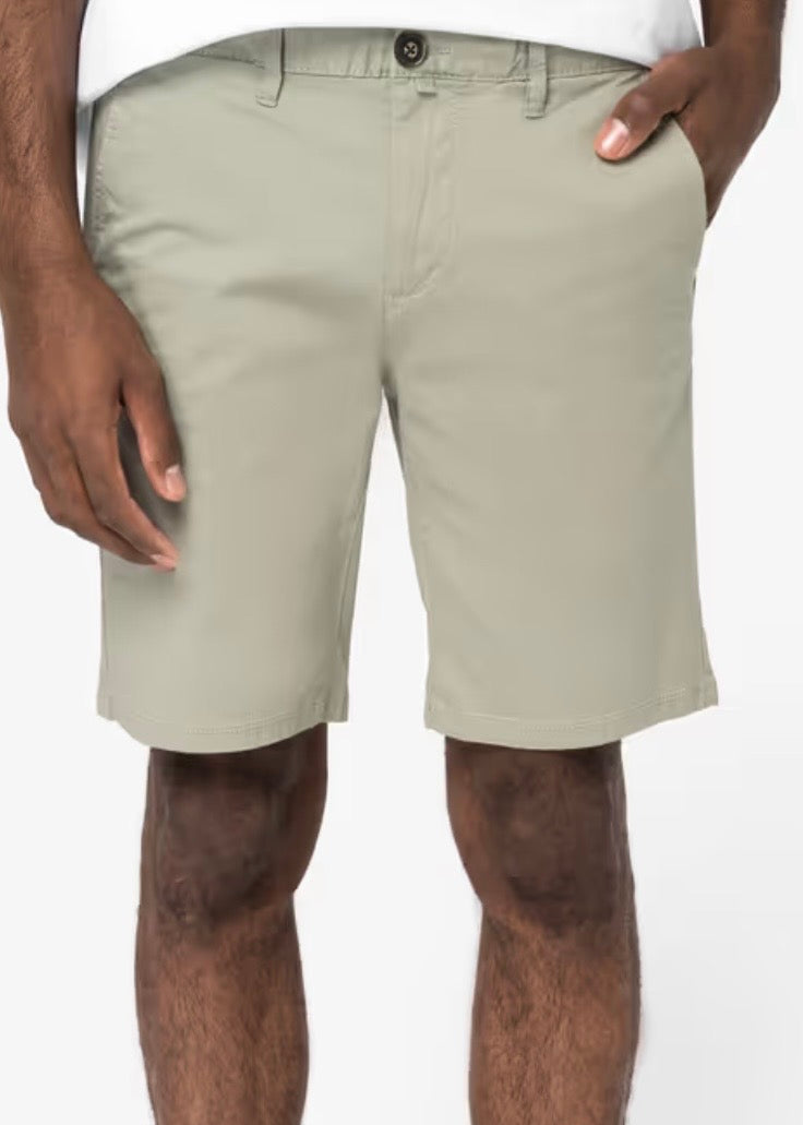 A man wearing almond green coloured chino shorts, with a recycled button, recycled metal zip, belt loops and side pockets, one hand is in a pocket, shown from the waist down against a neutral studio background. The shorts come down to just above his knees.