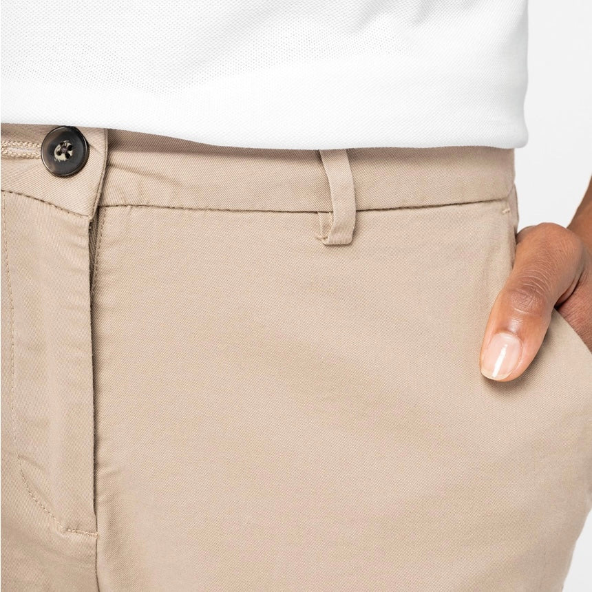 Close-up of sand dune coloured women's chino shorts worn with a white top, showing the waistband, recycled button, belt loops, and side pocket with a hand resting casually in it.