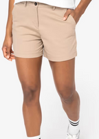 A woman wearing sand dune coloured chino shorts with a white top, white socks, and grey sneakers, shown from the waist down against a neutral studio background.