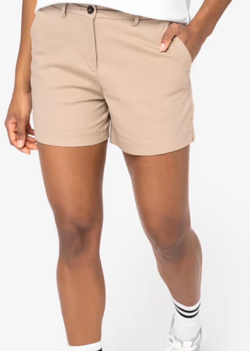 A woman wearing sand dune coloured chino shorts with a white top, white socks, and grey sneakers, shown from the waist down against a neutral studio background.
